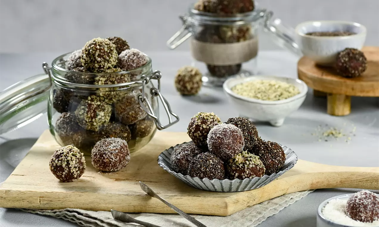 Boulettes rondes chocolatées recouvertes de noix de coco et graines, présentées dans un pot en verre et sur une petite assiette métallique