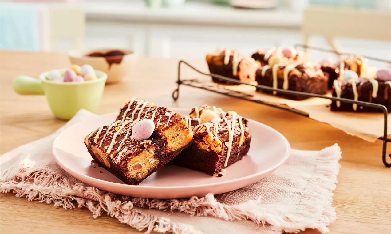 Brownies moelleux couleur cacao ornés de mini-œufs pastel et de filaments de chocolat blanc sur une assiette rose posée sur un tissu