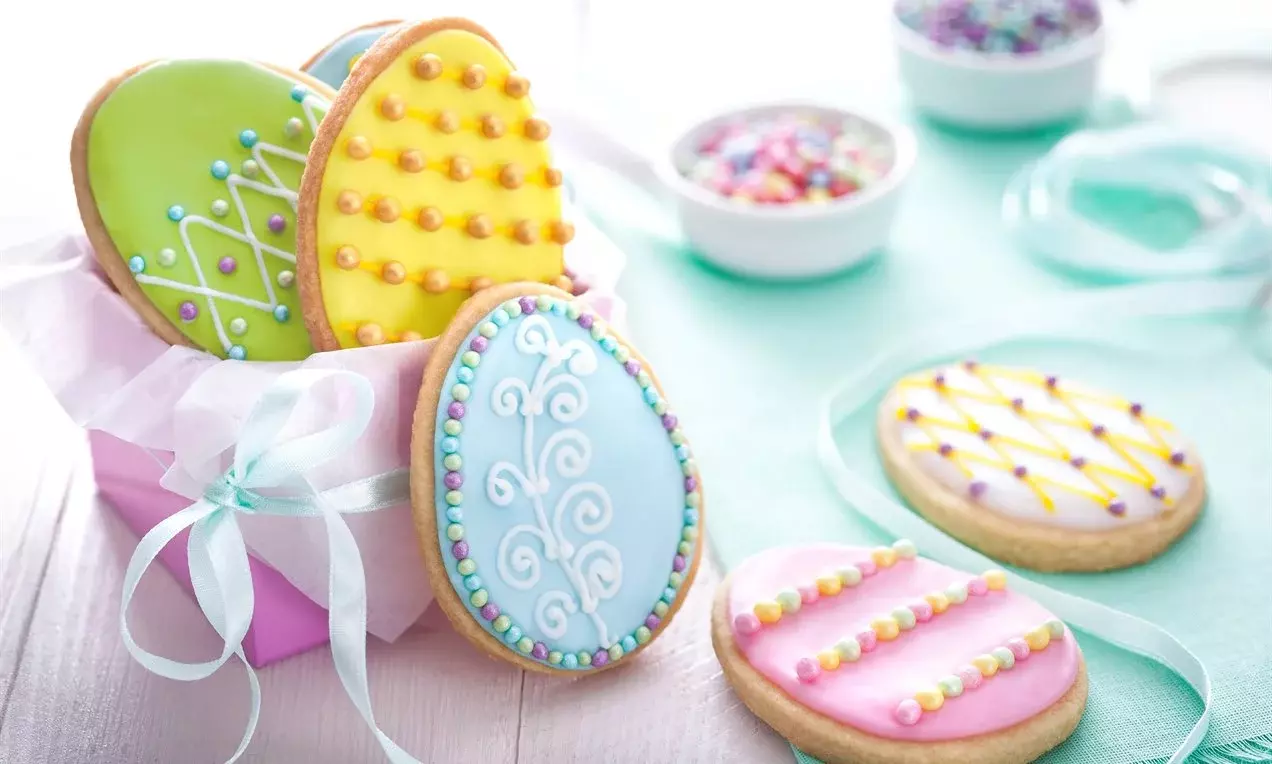Biscuits ovales décorés de glaçage pastel et motifs perlés dans une boîte rose nouée d’un ruban, accompagnés de petites coupelles