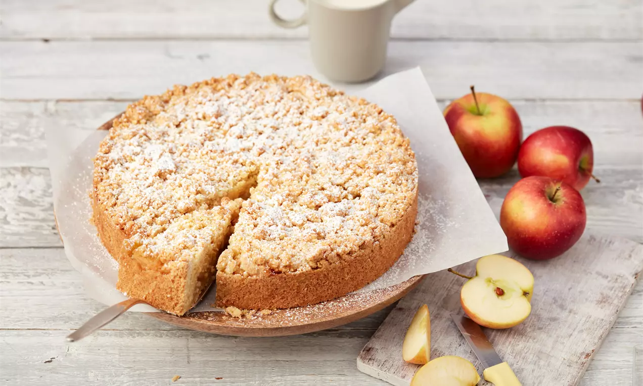 Gâteau streusel doré et moelleux surmonté de crumble poudré de sucre glace, entouré de pommes rouges et de tranches fraîches sur bois