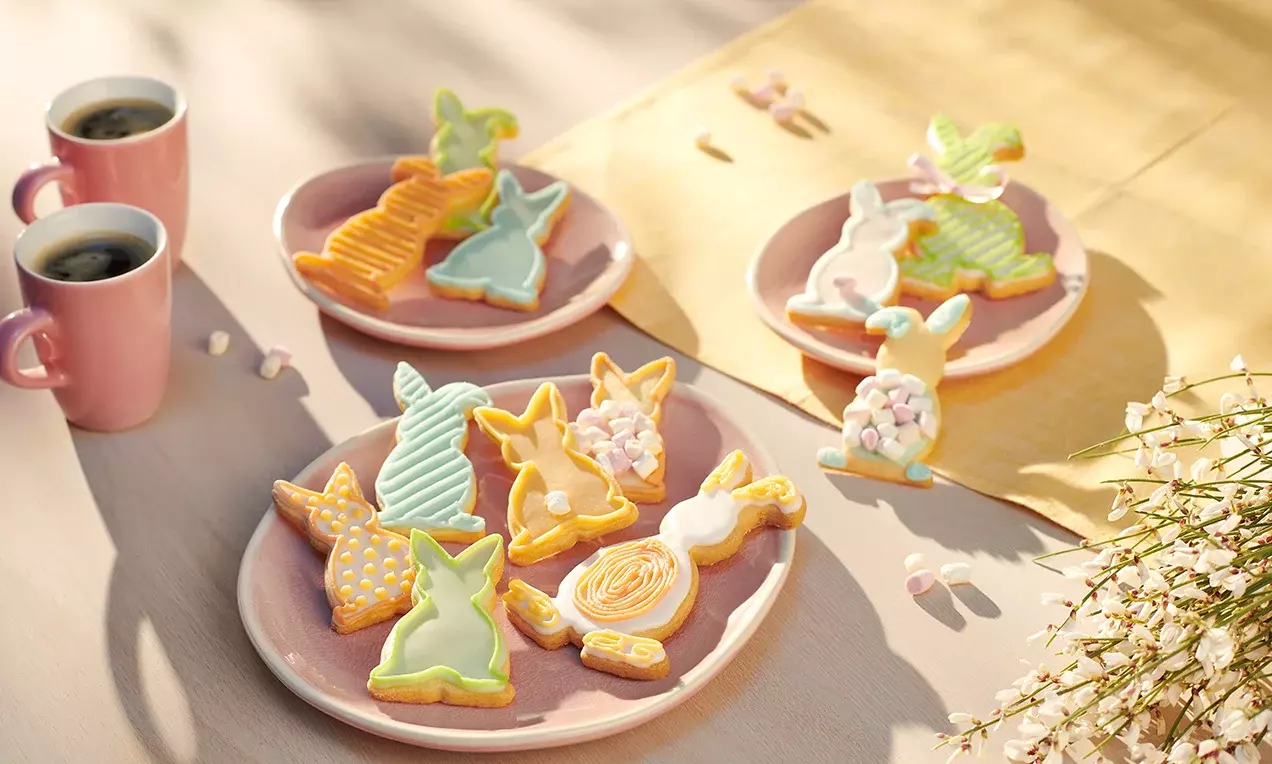 Biscuits en forme de lapins aux glaçages pastel orange, jaune et vert, disposés sur des assiettes roses près de tasses de café et fleurs blanches