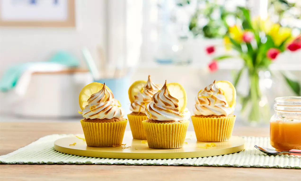 Petits gâteaux au citron jaune vif dans des caissettes dorées, surmontés de meringue dorée et de tranches de citron sur un plateau jaune