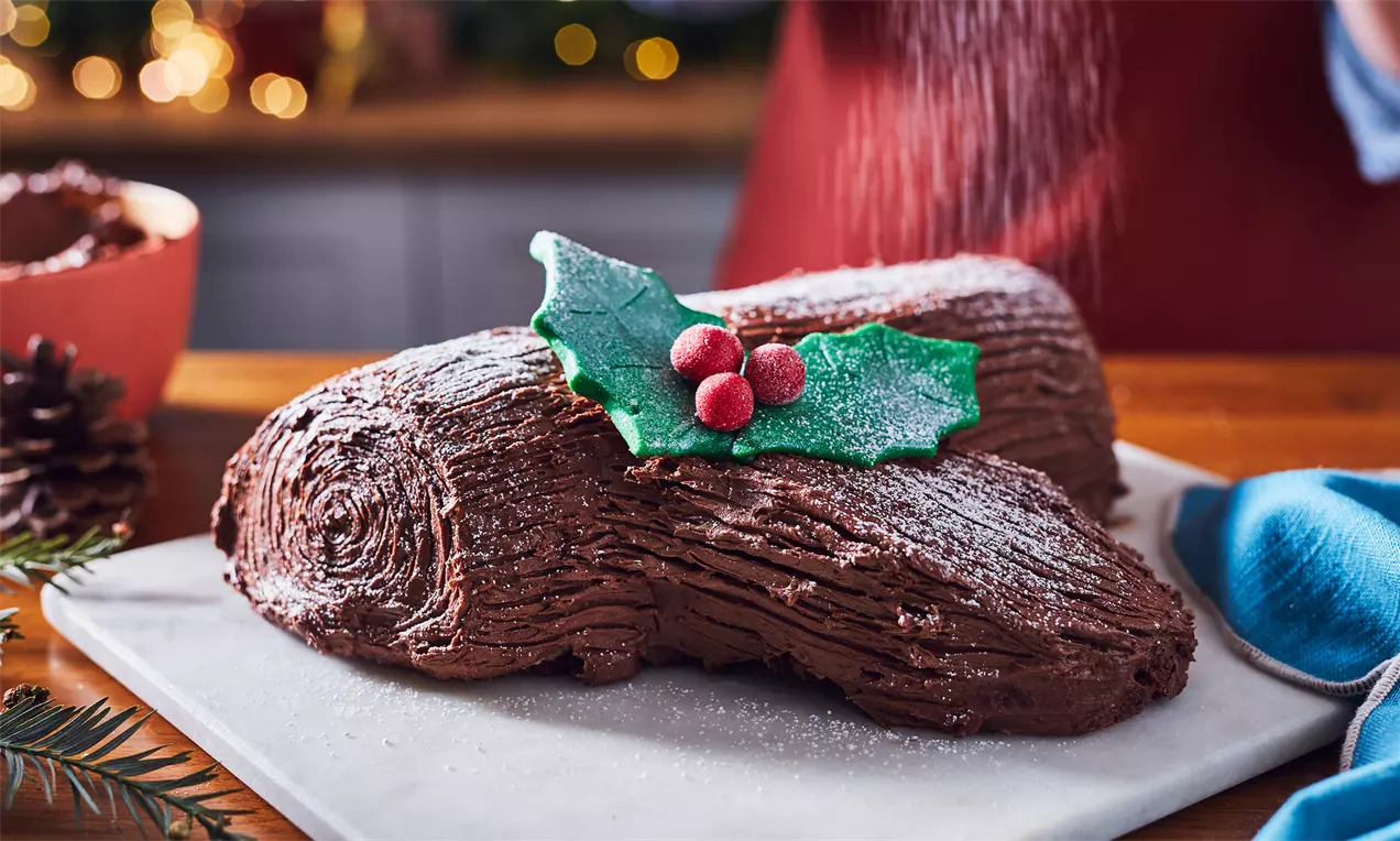 Bûche au chocolat brun texturée façon écorce, décorée de feuilles vertes en pâte d’amande et baies rouges sur une planche blanche