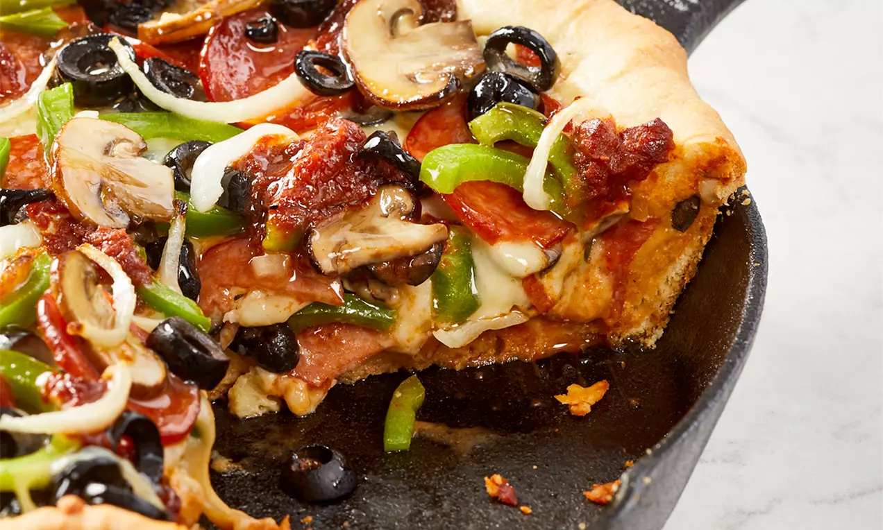 Pizza à pâte épaisse dorée garnie de fromage fondu, champignons, olives noires, poivrons verts et rondelles de pepperoni dans une poêle en fonte
