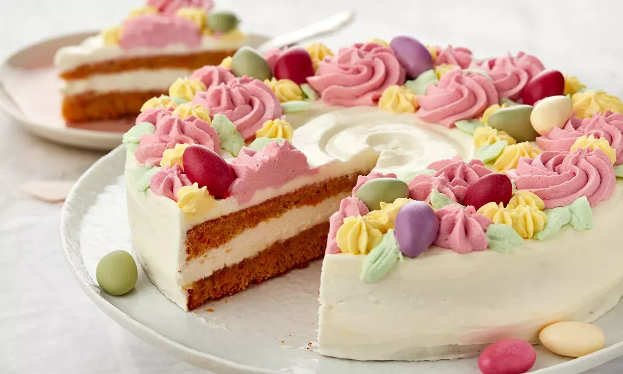 Gâteau rond à couches alternées de biscuit doré et crème blanche, décoré de rosaces pastel et d'œufs colorés sur une assiette blanche